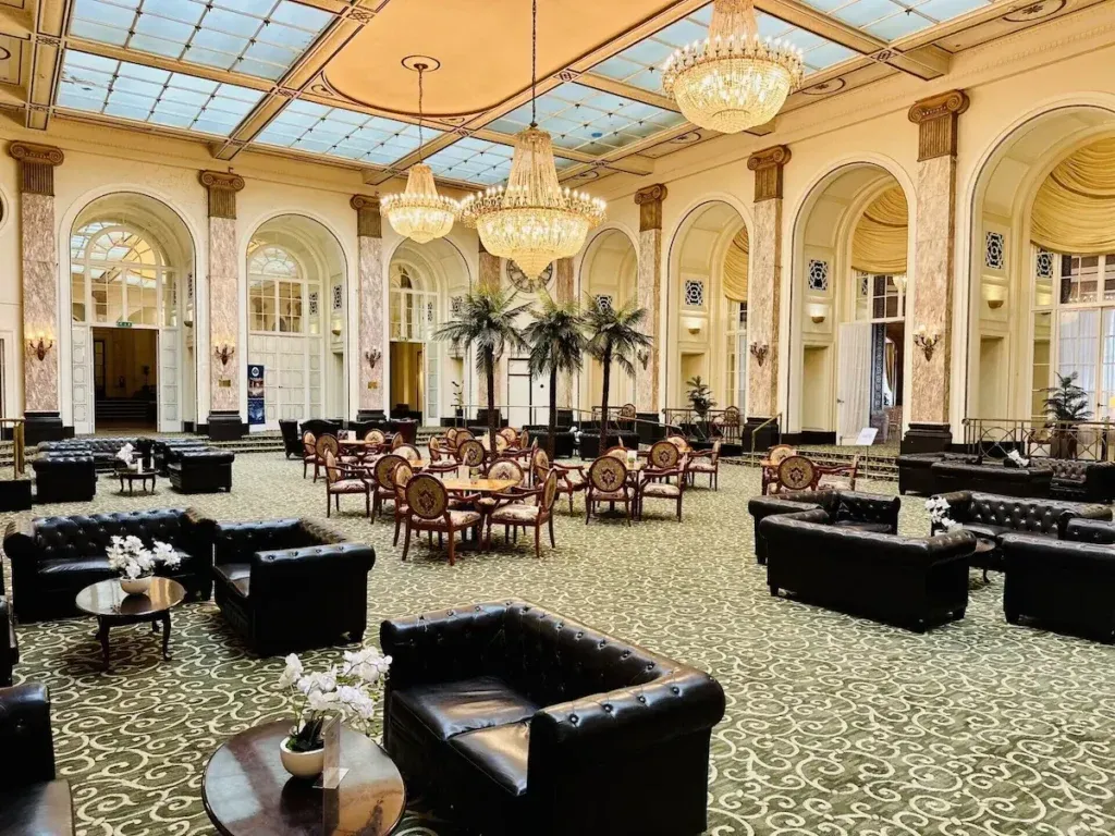 Grand lounge at Adelphi Hotel with a glass ceiling, crystal chandeliers, marble columns, arched doorways, black leather chesterfield seating and round tables with chairs