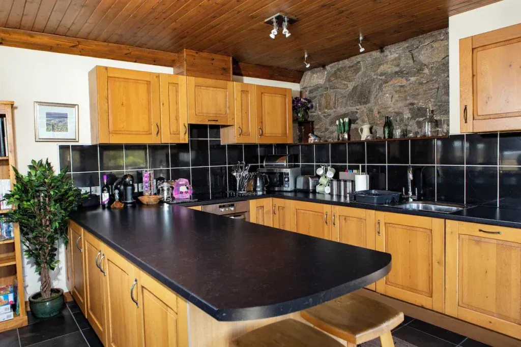Wooden kitchen cabinets, black worktops and a breakfast peninsula beside an exposed stone wall at An Taigh Clach