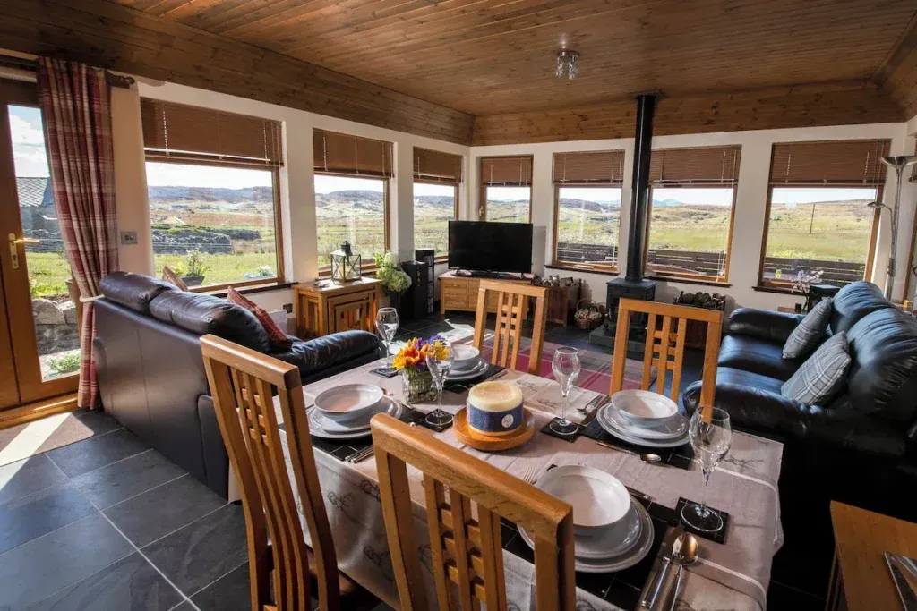 Dining table set for four beside leather sofas and a stove, with wide landscape views through the windows at An Taigh Clach