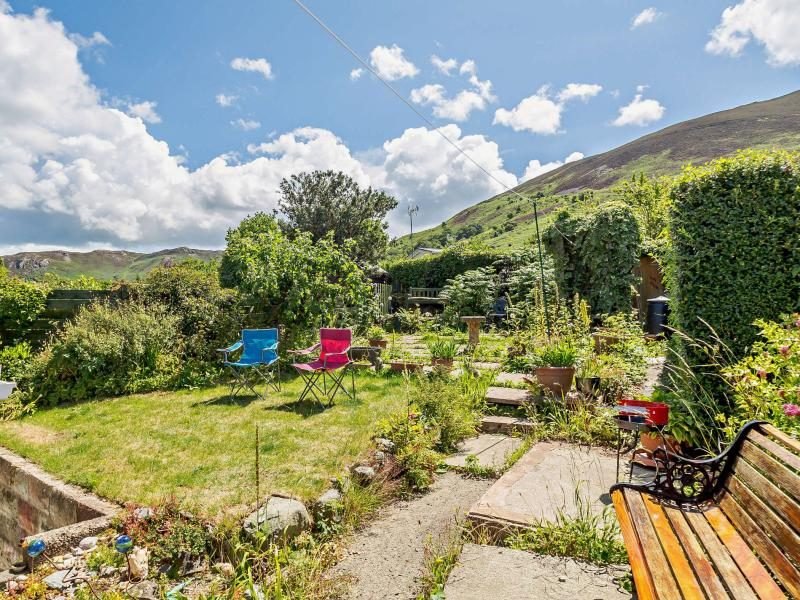 Garden with lawn, flowering borders, a bench and colourful folding chairs beneath a green hillside