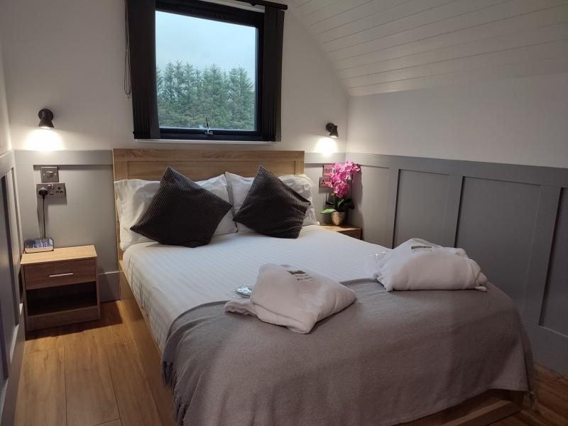 Double bed with grey throw and folded white bathrobes, bedside tables, wall lights and a window looking onto trees