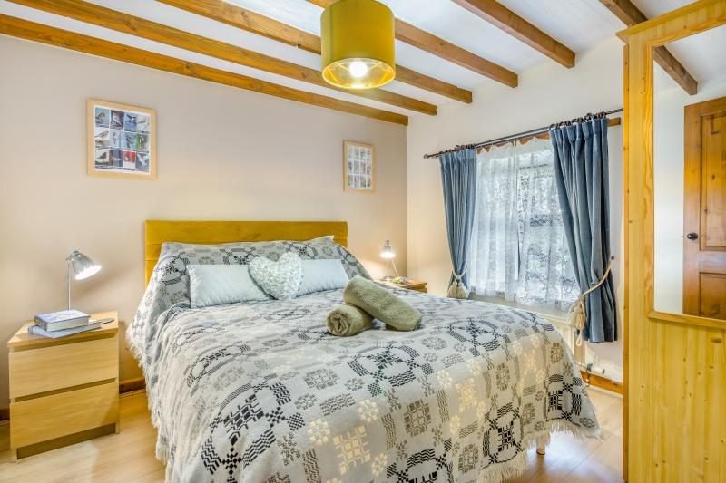 Double bedroom with exposed ceiling beams, a wooden bed with a woven patterned bedspread, blue curtains and bedside lamps