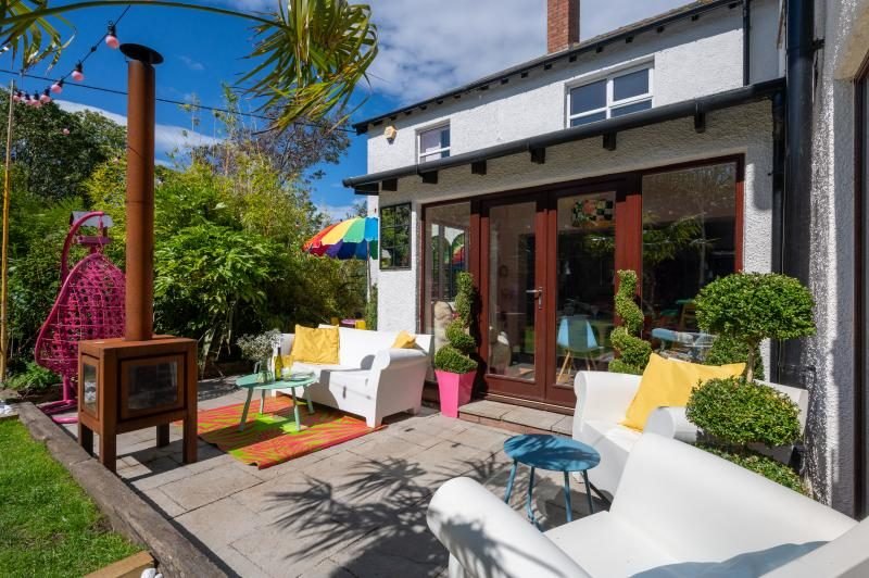 Colourful Cinema House near Craster, Northumberland 2 Paved patio with white sofas, a rust-coloured outdoor stove, pink hanging chair and a rainbow parasol