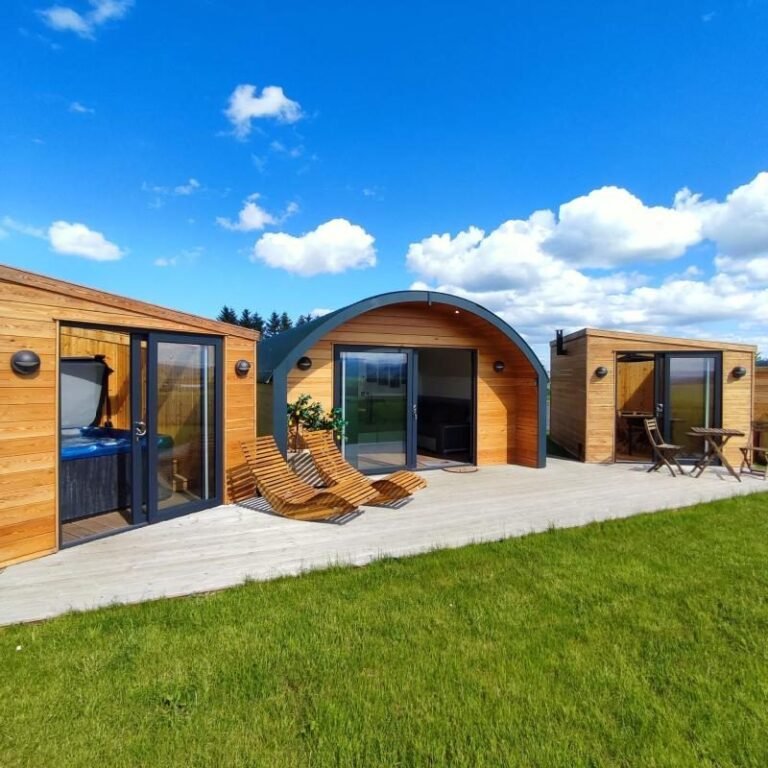 Timber glamping pod with curved roof between a glass-fronted hot tub house and a BBQ hut, with sun loungers on decking under a blue sky