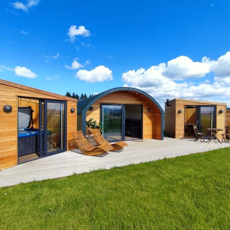 Timber glamping pod with curved roof between a glass-fronted hot tub house and a BBQ hut, with sun loungers on decking under a blue sky