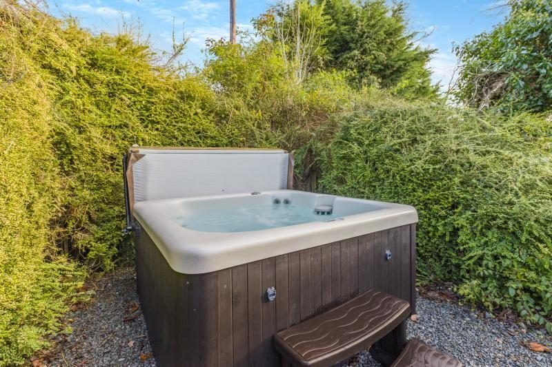 Hot tub with timber-style panels and steps on a gravel area, surrounded by tall green hedges