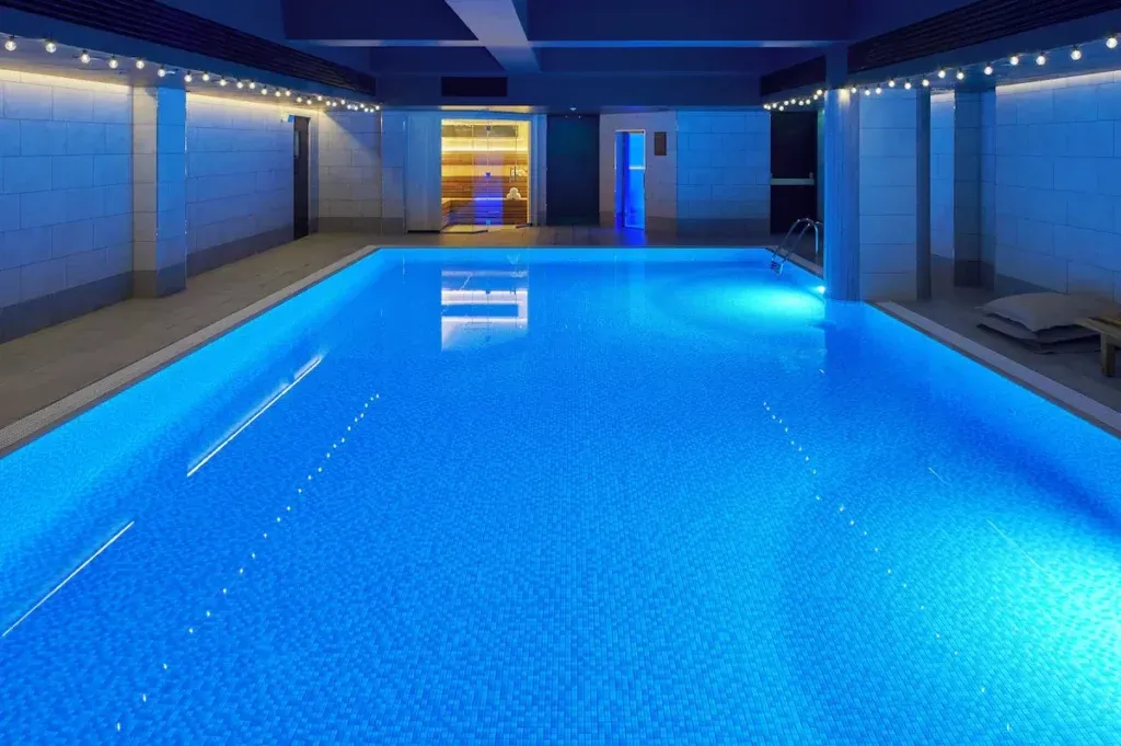 Blue-lit indoor swimming pool with tiled walls and poolside loungers at Kimpton Charlotte Square
