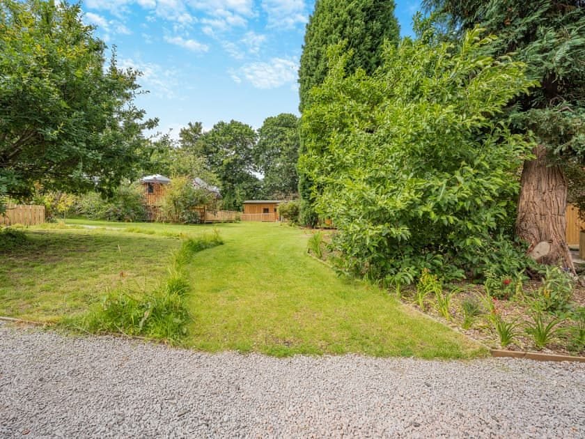 Lawned garden with mature trees and shrubs, a gravel drive in the foreground and timber lodges in the distance