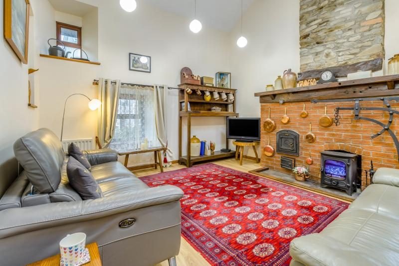 Living room with grey leather sofas, a red patterned rug and a woodburner set into a brick fireplace with copper pans on the wall