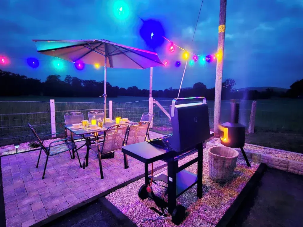 Patio at dusk with a set dining table and parasol, a gas barbecue and a freestanding fire pit, overlooking open fields under colourful string lights
