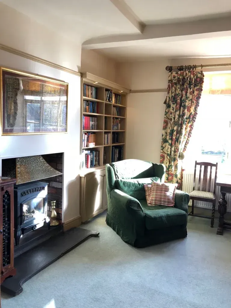 Green armchair beside a stove, bookshelves and patterned curtains in the sitting room