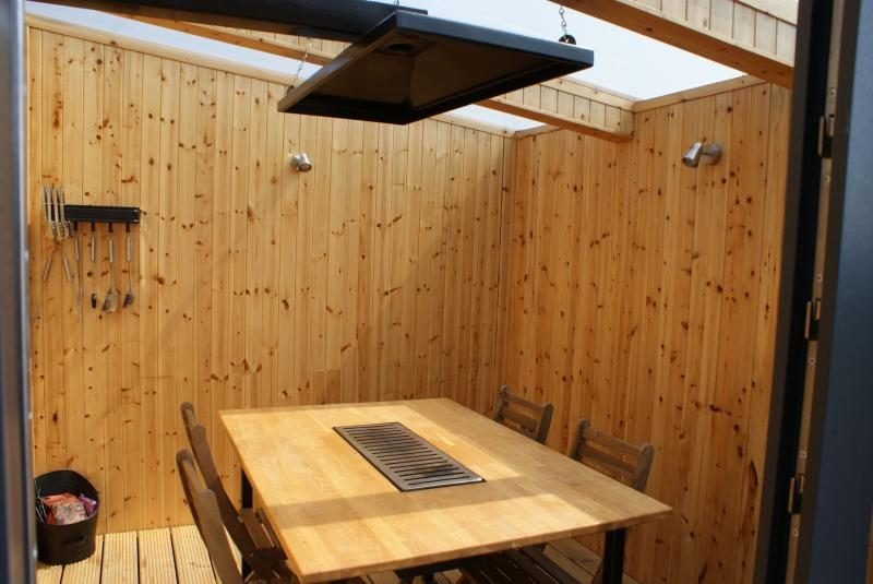 Timber-clad BBQ hut with a dining table with built-in grill, extractor hood above, wooden chairs and a rail of cooking utensils