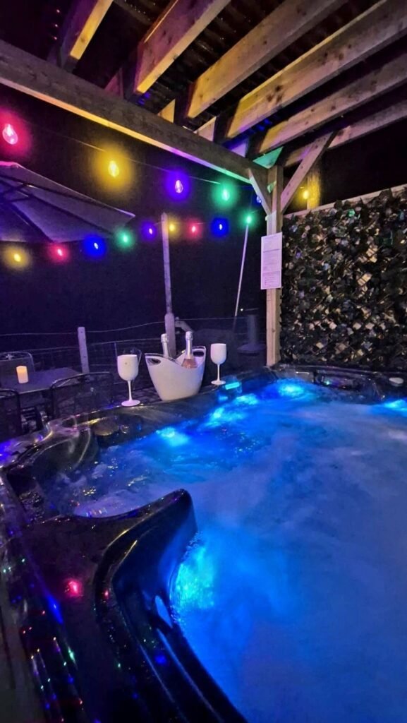 Illuminated hot tub at night under timber beams, with colourful string lights, a bucket with two bottles and two glasses, and a patio table behind