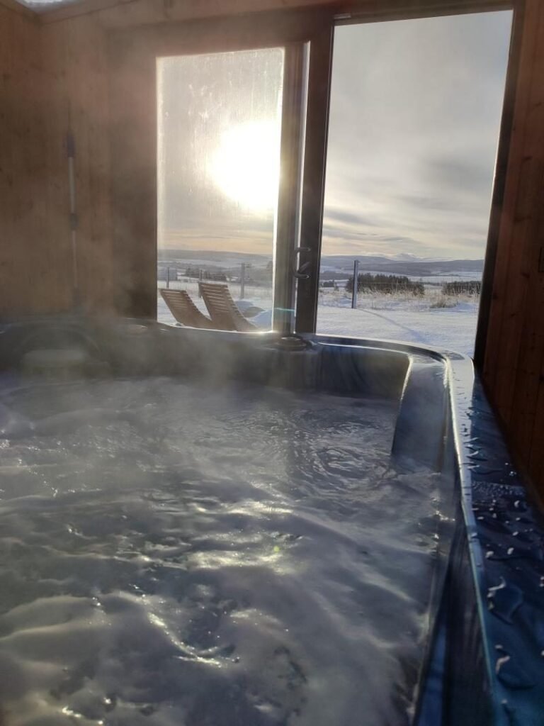 Steaming hot tub inside a timber hot tub house with glass doors opening onto sun loungers and a snowy landscape