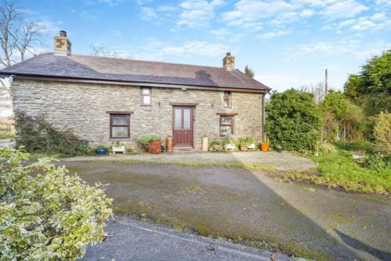 Stone-built cottage with a slate roof, two chimneys and a red front door, with potted plants along the front and a drive in the foreground