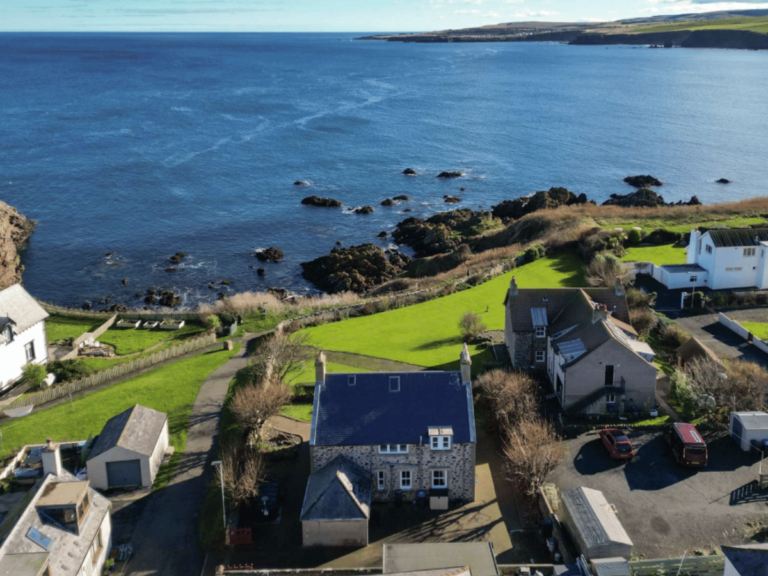 Aerial view of stone houses and gardens beside the rocky coastline and blue sea at St Abbs in the Scottish Borders