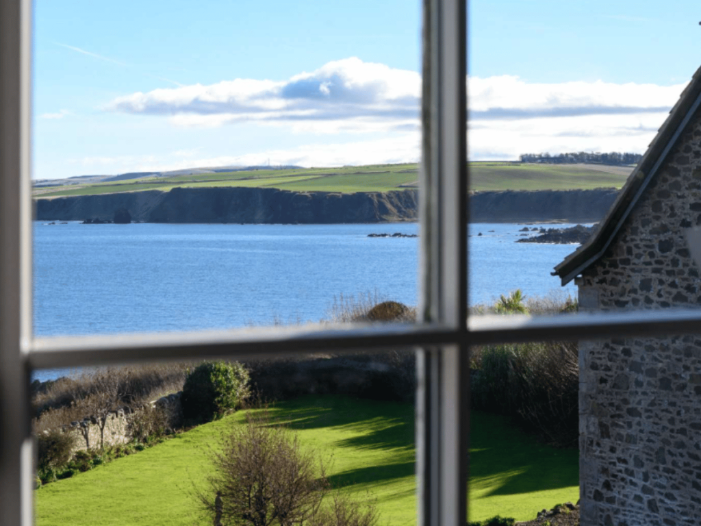 View through a window overlooking a green garden, blue sea and coastal cliffs at The Beacon in St Abbs