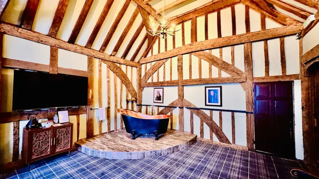 Freestanding bath on a raised tiled platform in a suite at The Ostrich Inn with exposed timbers and a wall-mounted television