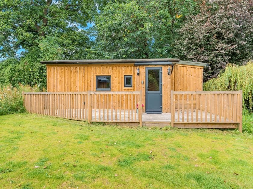 Timber-clad single-storey lodge with a grey door, wooden deck and railings set on a lawn surrounded by trees
