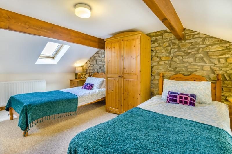 Twin bedroom with a sloping beamed ceiling, an exposed stone wall, two single beds with teal throws, a pine wardrobe and a skylight